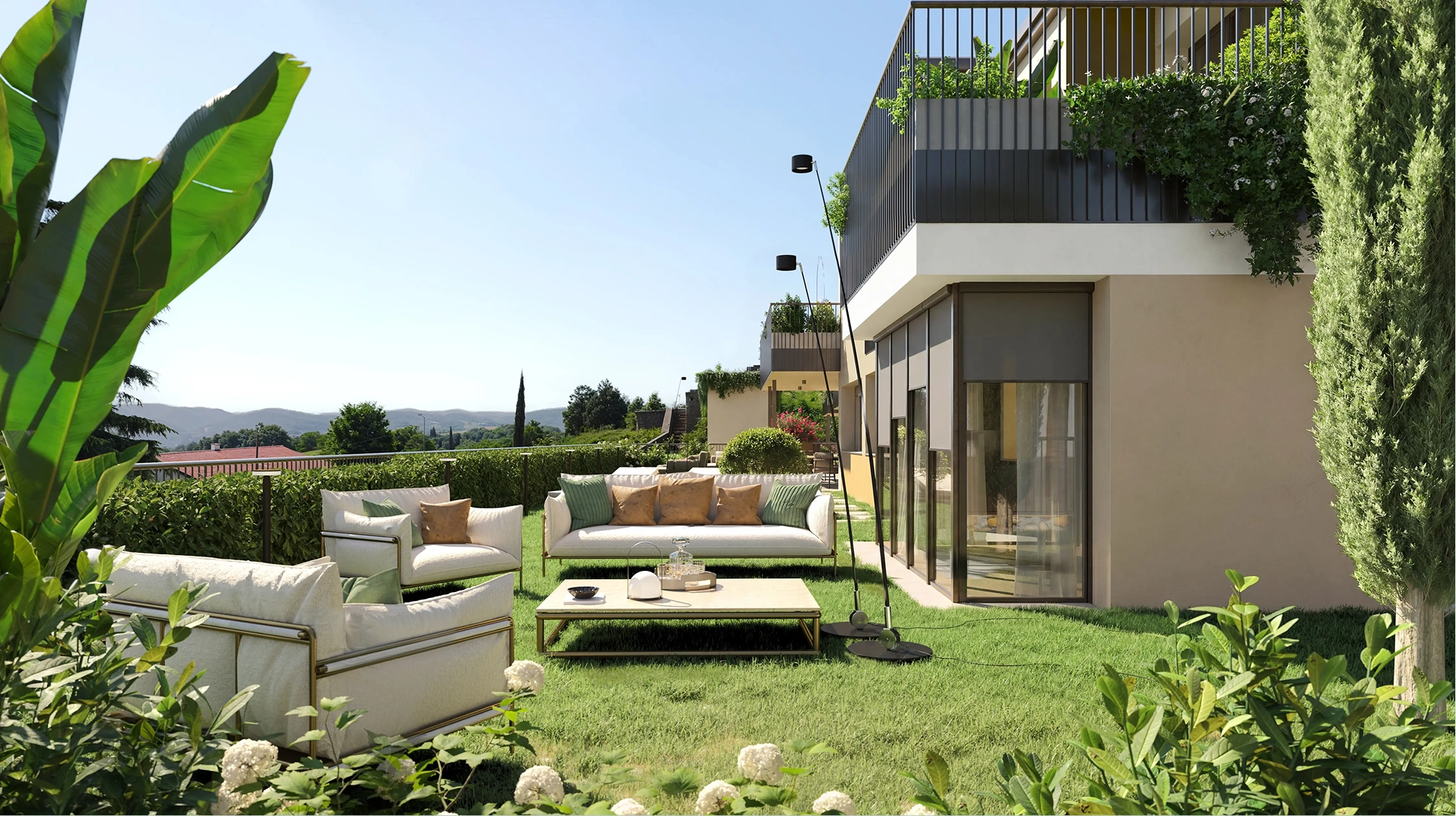 View from a balcony overlooking an outdoor pool, sun loungers, green lawn, and lush trees with distant hills under a clear sky.
